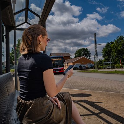 Frau sitzt mit ihrem Handy in der Hand an einer Bushaltestelle